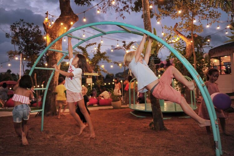 Children play on a curved metal climbing frame in an outdoor park in Parklife Bali at twilight, with strings of lights adding a cozy atmosphere.