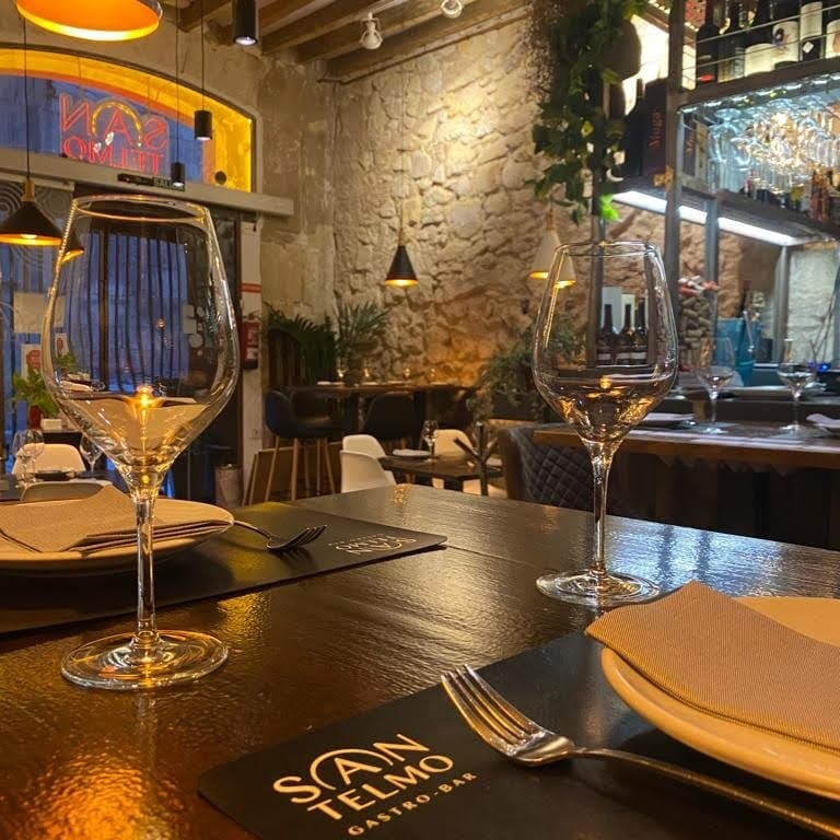 Mesa elegante y dispuesta en el interior de San Telmo Gastrobar, con copas de vino perfectamente alineadas. Ambiente cálido y sofisticado, que hace resaltar las paredes de piedra natural y detalles en madera. La atmósfera invita a disfrutar de una experiencia gastronómica íntima y acogedora en pleno Casco Antiguo de Alicante.