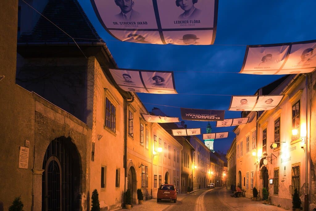 Esti kép az Elfeledett Soproniak kiállítás utcai napvitorláiról. Balra az Ó-zsinagóga bejárata.