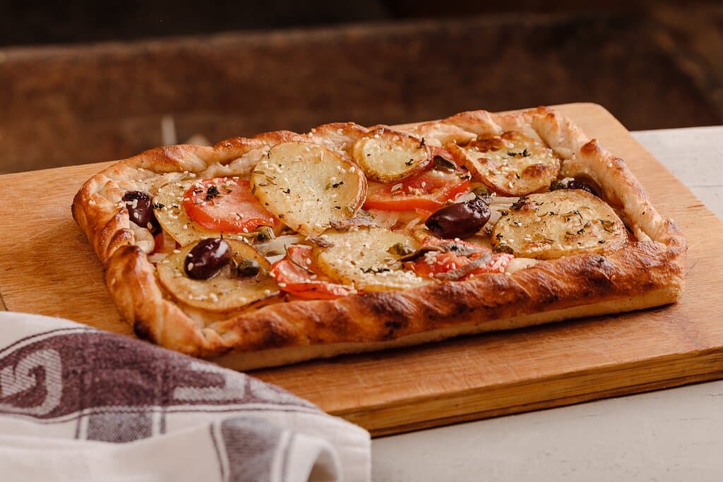 Karmni s-Sultana
Maltese bread dough topped with thin sliced potatoes, tomatoes, anchovies, onions, capers, olives, fresh mint, fennel seeds and sesame seeds.