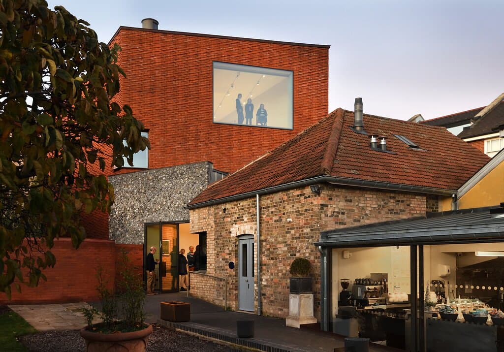 An image taken from within the gardens at Gainsborough's House. It shows The Watering Place cafe along with the back entrance to the new art galleries. There are visitors looking out of the large, Landscape Studio window. The 400 year old Mulberry Tree is also in shot. 