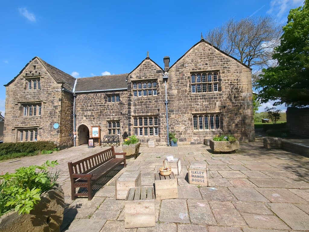 The front of Ilkley Manor House in summer. There are chalks and toys on Manor House boxes. The blue plaque is to the left of the front door, which is open. On the right in the distance is one of the interpretation stones about life in Roman Ilkley. 