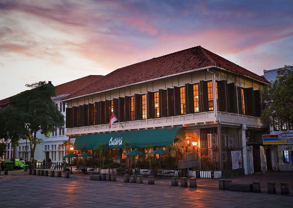 Café Batavia at twilight. Nestled in a historic corner, the restaurant's colonial architecture stands majestically against a canvas of the setting sun. Its warm, glowing windows invite passersby to partake in an experience that promises a blend of cultural richness and culinary delight.