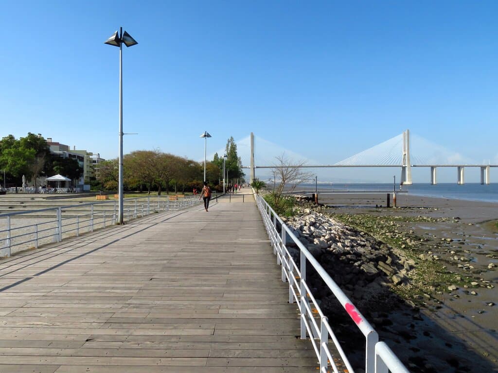 Passerella in legno che attraversa un canale; il ponte Vasco da Gama domina sempre l'orizzonte a nord