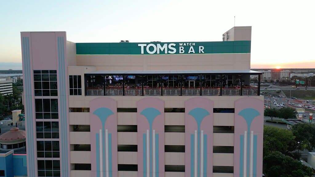 Tom’s Watch Bar from above, highlighting its vibrant pastel-colored exterior and spacious outdoor patio with large screens. The iconic sign stands out against the sunset and city backdrop, making it a standout spot for sports and fun. 