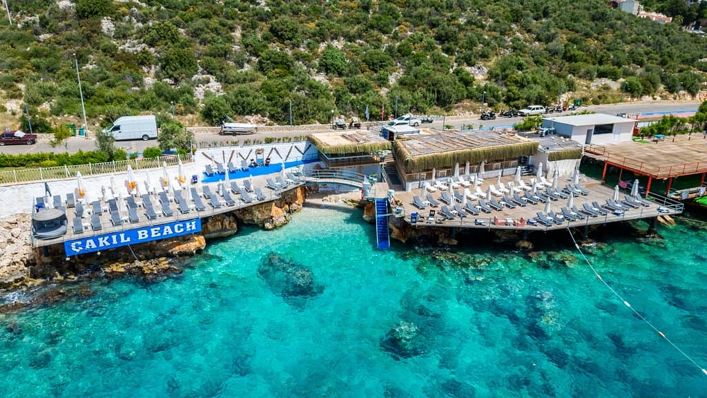 The Cool Breeze and Warm Welcome of Kalkan: Welcome to Çakıl Beach! Nestled in one of Kalkan’s most beautiful coves, enjoy the sea your way — whether by walking in from our natural shoreline or using our stair-access deep entrance. Refreshing waters, tranquility, and nature await you.  Relax on our spacious sunbathing decks or unwind in the comfort of our private cabanas. Our freshly made cocktails and delicious dishes are always ready to elevate your beach day.  Easily accessible from Kalkan to