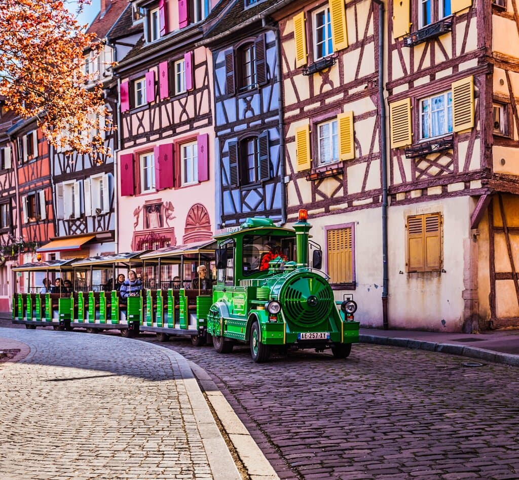 Le petit train vert de Colmar longe les rangées de maisons pittoresques qui font la renommée de la vieille ville. Leurs façades à colombages, peintes dans des tons vifs de rouge, bleu, jaune et vert, créent un décor vivant et chaleureux, digne d’un conte de fées alsacien. Tandis que le train avance paisiblement, les passagers sont plongés dans une atmosphère joyeuse et charmante, au cœur de l’âme authentique de Colmar.