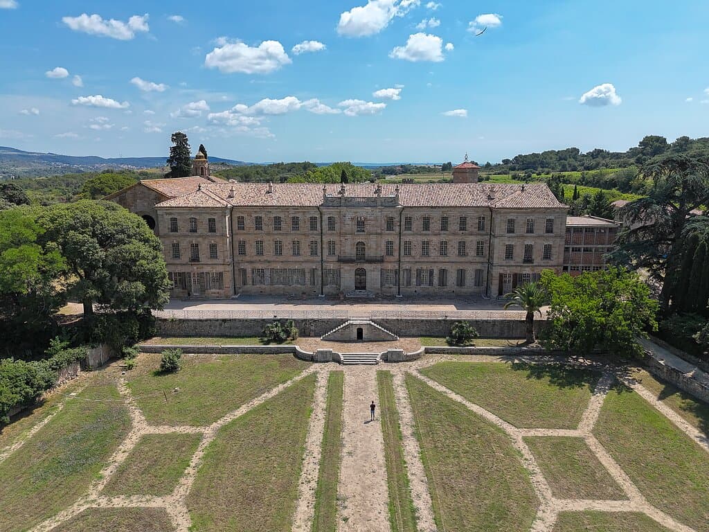 Sous l'azur éclatant du Sud, le Château-Abbaye de Cassan se dresse avec une noblesse tranquille, témoin de siècles d'histoire gravés dans sa pierre blonde. Cette vue aérienne nous offre un face-à-face saisissant avec la symétrie parfaite de sa façade néoclassique, dont chaque fenêtre semble garder le secret d'une époque révolue.