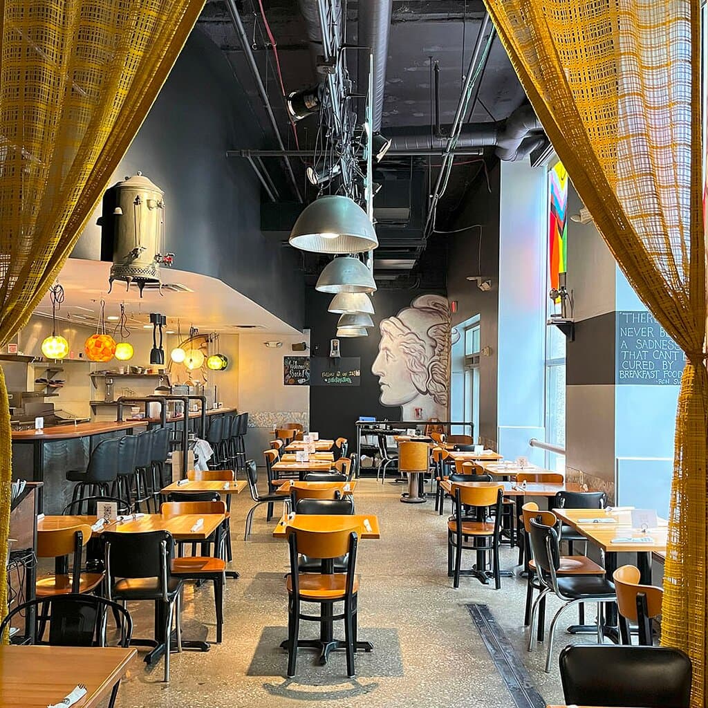 A peek into the heart of Dime Store Detroit—our bright, eclectic dining room as seen from the gold-curtained nook. This view shows off our vintage hanging light fixtures, colorful globe lights over the counter, and the iconic Mercury dime mural at the far end. Even without guests, the space glows with warmth and character, reflecting the inviting, offbeat energy of one of downtown Detroit’s favorite brunch spots.